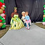 balloon_arch, child, toddler, princess_costume, roller_skates, balloons, costume_party, smiling, outdoor, concrete_path, black_backdrop, green_gloves, yellow_dress, pink_tutu, curly_hair, happy, people, event, celebration, colorful