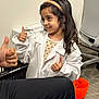 box, carpet, casual, child, clothing, expression, happy, headband, human_hand, indoor, interaction, long_hair, office_chair, orange_bucket, person, positive_gesture, smiling, thumbs_up, white_lab_coat, young_girl
