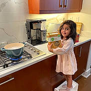 Aadya joined the competition — help win amazing prizes! child, girl, kitchen, stove, pot, pasta_box, step_stool, socks, dress, smile, cooking, countertop, cabinet, appliance, modern, indoor, happy, food, young_child, curly_hair