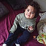 toddler, child, curly_hair, smiling, sitting, bed, blanket, pillow, hoodie, long_pants, white_socks, indoors, waving_hand, face, person, cozy, soft_toys, maroon_bedspread, casual_clothing, cute