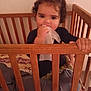toddler, child, crib, wooden_crib, curly_hair, baby, blanket, bedding, indoor, person, face, hand, clothing, curious, innocent, holding, soft, wall, furniture, standing
