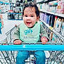 toddler, child, shopping_cart, store, aisle, shelves, products, baby, curly_hair, green_bib, blue_jeans, happy, smiling, hands, shopping, retail, indoor, person, casual_clothing, bracelet