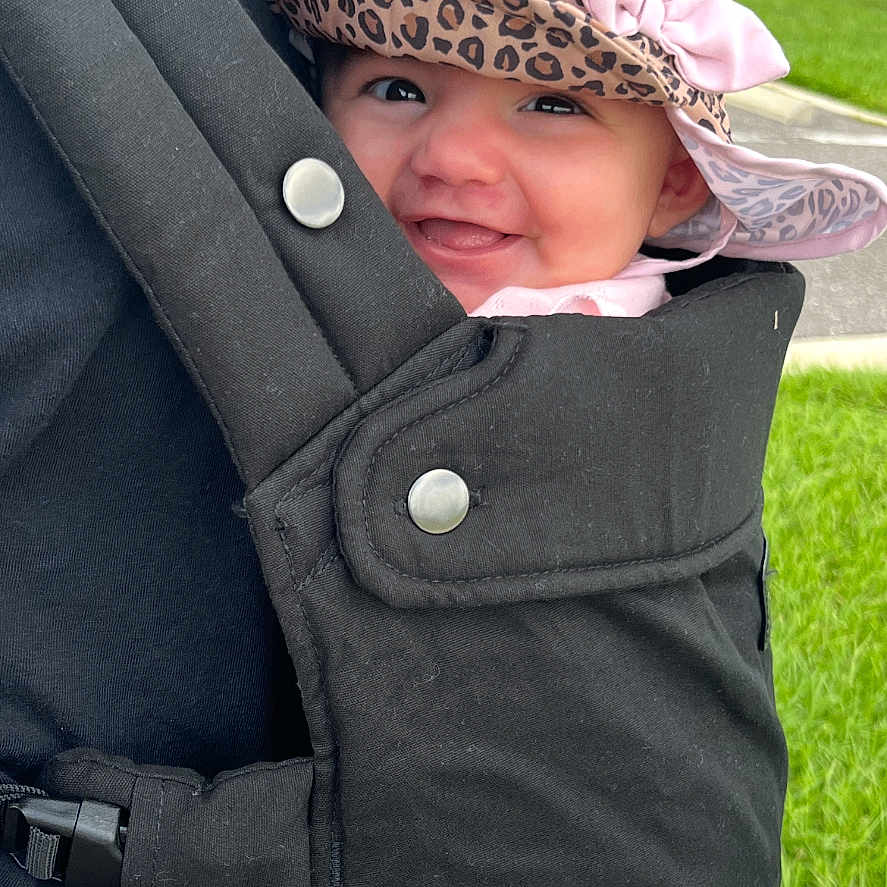 Elizabeth is registered to the contest to win money with this photo: adult, baby, baby_carrier, black_clothing, cute, daylight, grass, greenery, hand, happy, hat, infant, leopard_print, outdoor, park, person, pink_bow, side_view, smile, trees
