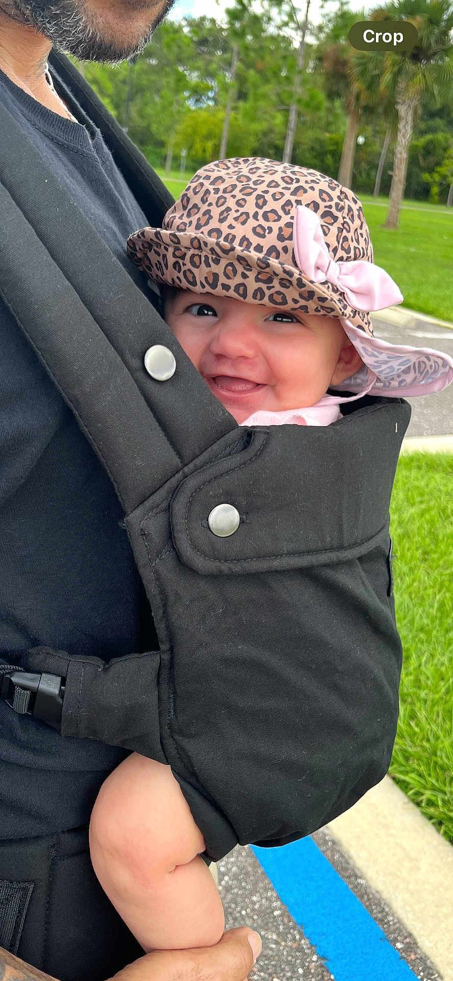 Elizabeth is registered to the contest to win money with this photo: baby, infant, smile, hat, leopard_print, pink_bow, baby_carrier, black_clothing, adult, hand, outdoor, greenery, trees, grass, side_view, happy, cute, person, park, daylight