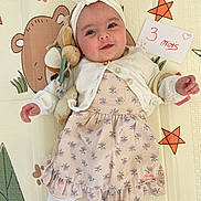 Thea participe au concours pour gagner de l'argent avec cette photo : animal_print, baby, cardigan, child, cute, dress, girl, handwritten_sign, headband, indoor, infant, lying_down, milestone, patterned_dress, playmat, smiling, soft_toys, stars, toy, white_clothing