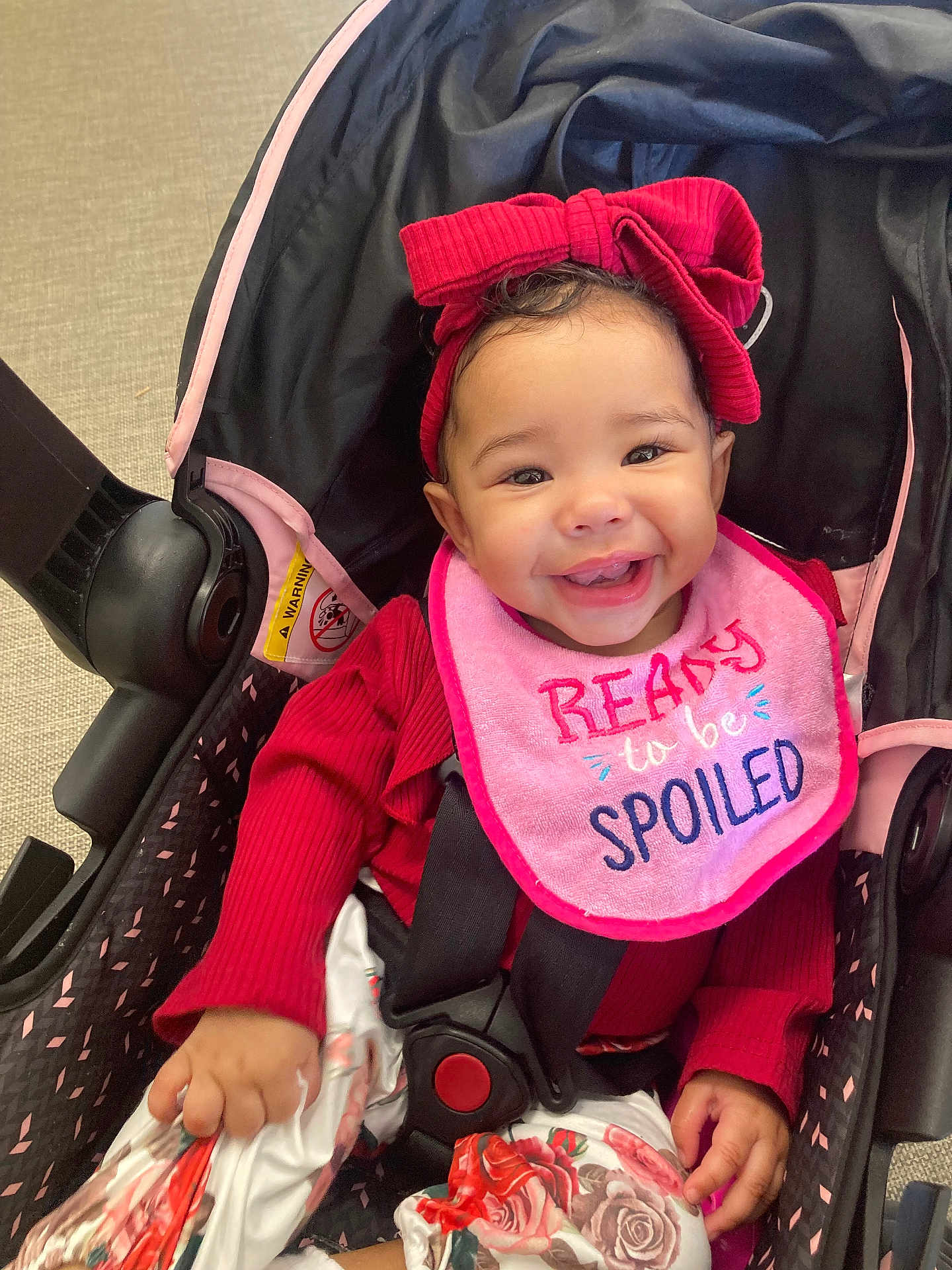 Sinayah is registered to the contest to win money with this photo: baby, baby_girl, car_seat, child, clothing, cute, face, floral_pants, happy, headband, indoors, infant, person, pink_bib, red_bow, red_outfit, seatbelt, sitting, smiling, stroller