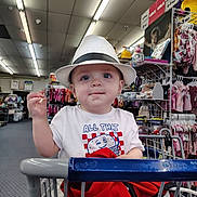 Noah is registered to the contest to win money with this photo: baby, child, shopping_cart, hat, fedora, clothing, store, retail, indoor, portrait, cute, blue_eyes, smile, infant, rack, baby_clothes, shopping, casual_wear, person, curious