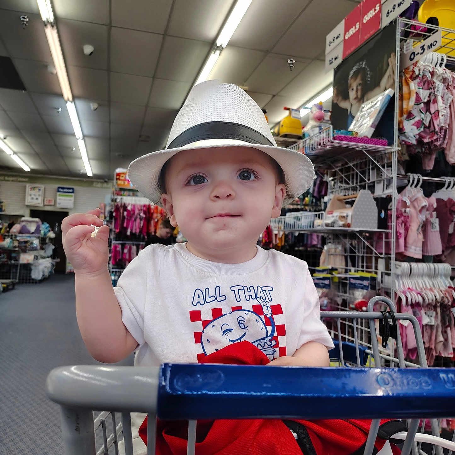 Noah is registered to the contest to win money with this photo: baby, child, shopping_cart, hat, fedora, clothing, store, retail, indoor, portrait, cute, blue_eyes, smile, infant, rack, baby_clothes, shopping, casual_wear, person, curious