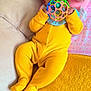 baby, blanket, bright_colors, child, closeup, colorful_toy, couch, cozy, curly_hair, cute, indoor, infant, pink_pillow, playful, portrait, relaxed, smiling_eyes, soft_texture, toy, yellow_onesie