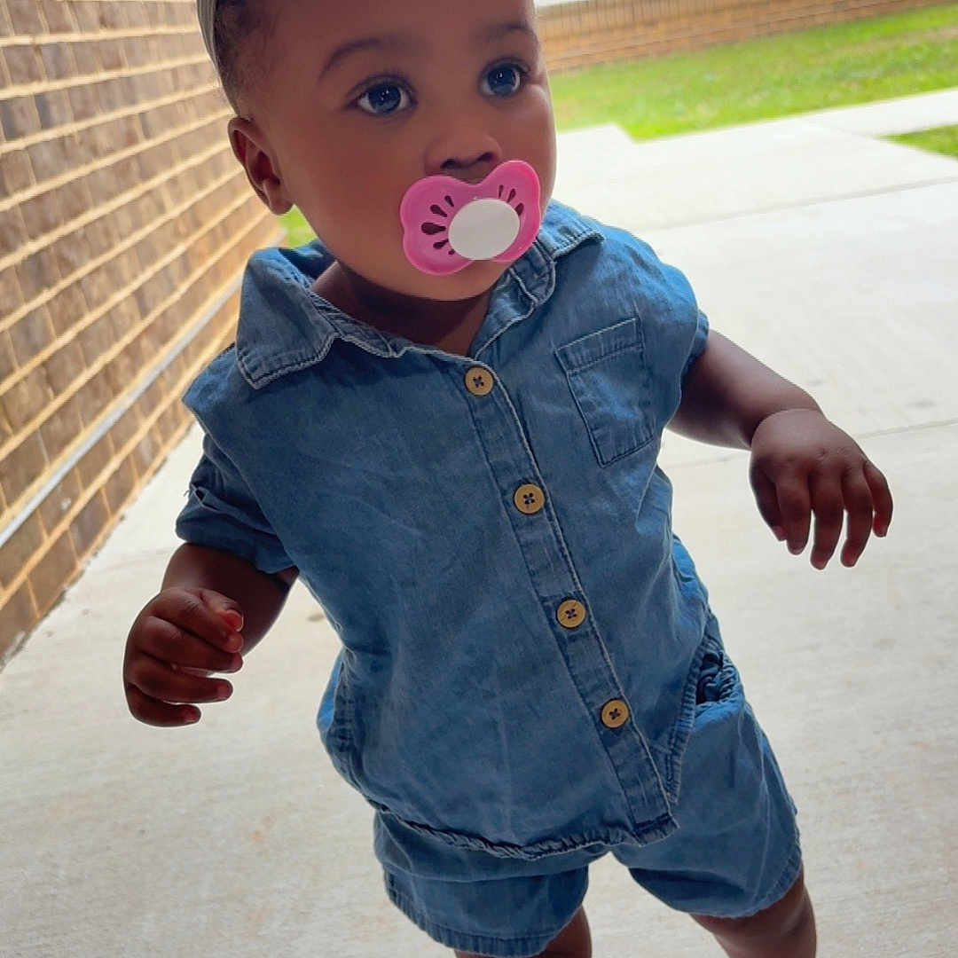 Kamorra is registered to the contest to win money with this photo: blue_outfit, bow, brick_wall, casual_clothing, child, concrete, curious, daylight, denim, footwear, headband, outdoor, pacifier, person, portrait, sandals, sidewalk, standing, toddler, young_child