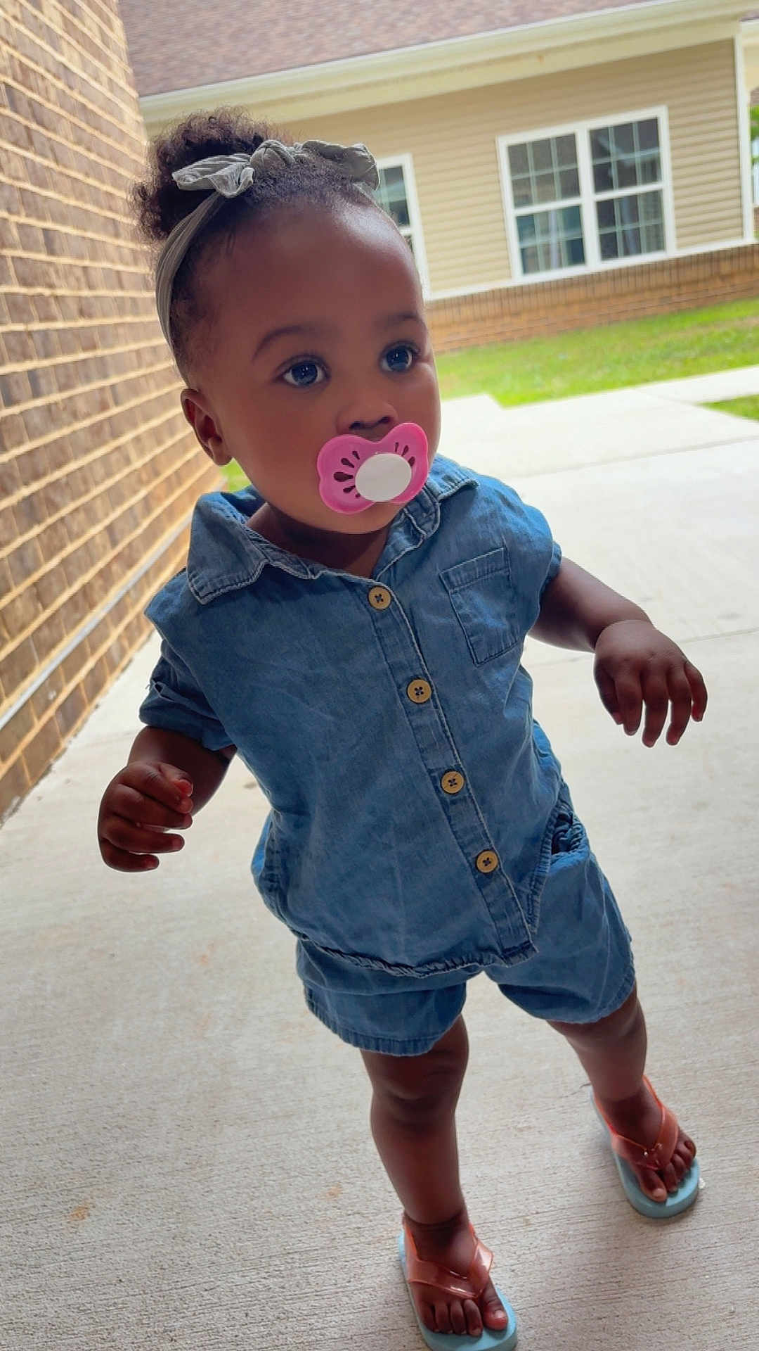 Kamorra is registered to the contest to win money with this photo: toddler, child, pacifier, denim, blue_outfit, sandals, headband, bow, outdoor, sidewalk, brick_wall, curious, standing, person, young_child, casual_clothing, daylight, portrait, footwear, concrete