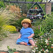 Robin joined the competition — help win amazing prizes! toddler, child, hat, blue_outfit, barefoot, garden, flowers, greenery, pathway, plants, outdoor, nature, sitting, smiling, sunny, daytime, pottery, plant_shelf, brick, leaves