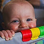 baby, child, face, smiling, playpen, colorful, hand, blue_eyes, indoor, person, adult, blurred_background, happy, infant, toys, mesh, grip, closeup, parent, home