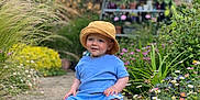 Robin is registered to the contest to win money with this photo: toddler, child, blue_clothing, bucket_hat, barefoot, garden, flowers, greenery, pathway, outdoor, plants, nature, smiling, sitting, daylight, pot_plants, rustic_shelf, colorful, cute, portrait