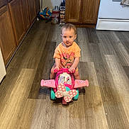 Skylar is registered to the contest to win money with this photo: toddler, child, toy, ride_on_toy, minnie_mouse, pink, kitchen, flooring, wooden_floor, cabinet, stove, appliance, childrens_toy, indoor, person, clothing, short_sleeve_shirt, smile, play, curious