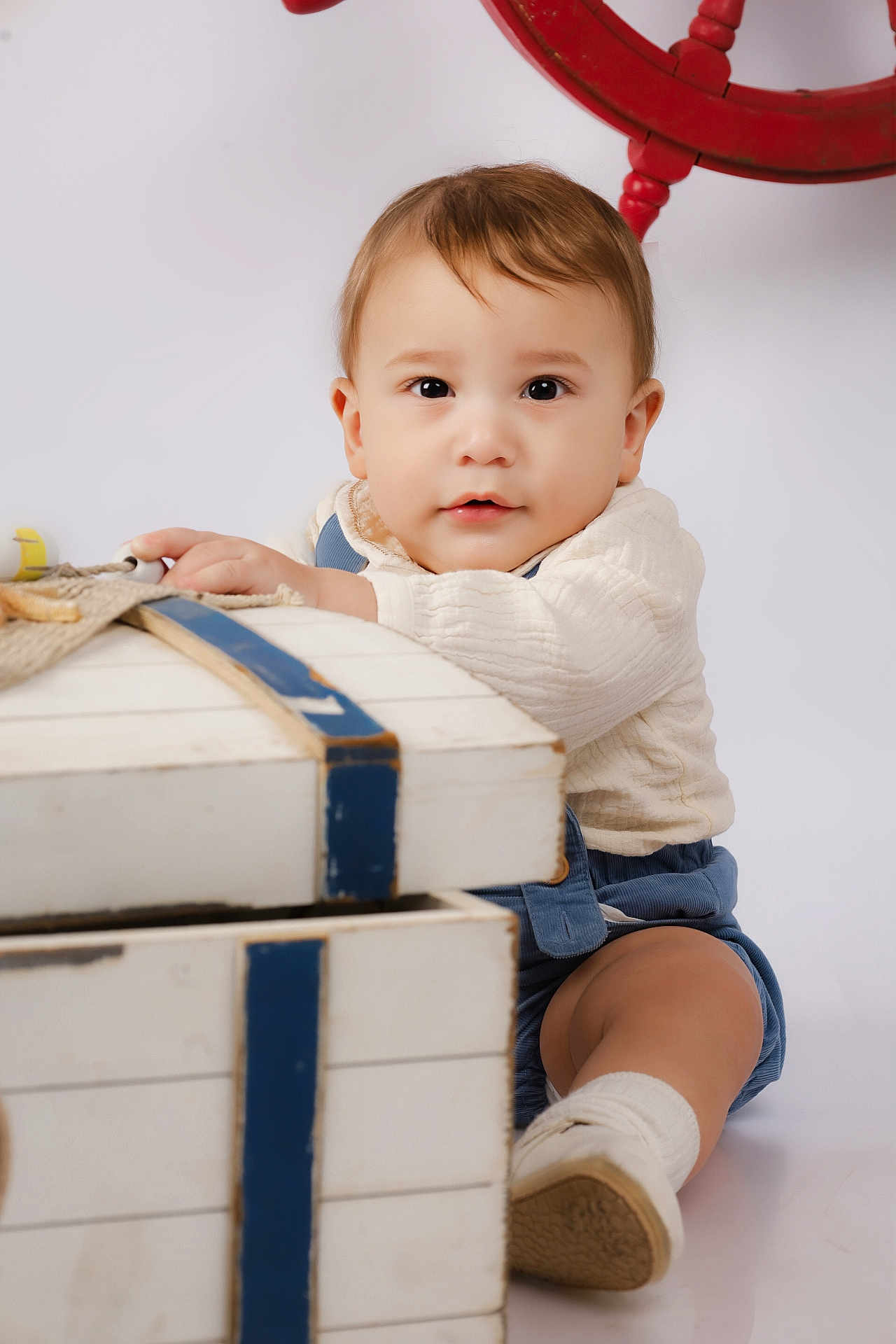 Thiago is registered to the contest to win money with this photo: baby, child, toddler, face, clothing, white_background, wooden_chest, blue_stripes, socks, shoes, indoor, portrait, cute, curious, sitting, hand, red_ship_wheel, studio_photo, soft_lighting, young_child