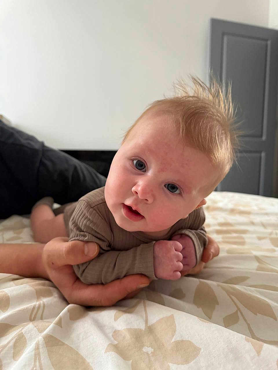 Yeva joined the competition — help win amazing prizes! baby, infant, hand, bed, bedspread, pattern, person, face, eyes, hair, skin, clothing, indoor, curious, lying_down, adult_hand, soft_light, closeup, portrait, expression