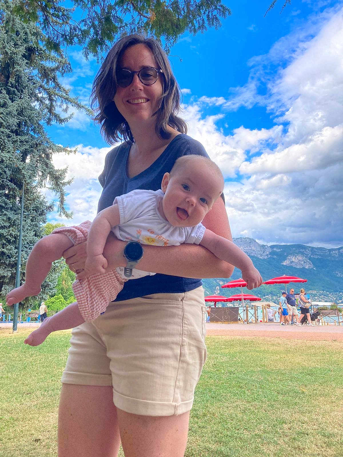 Anna participe au concours pour gagner de l'argent avec cette photo : woman, baby, child, outdoor, grass, sky, clouds, tree, glasses, shorts, tshirt, watch, red_umbrella, people, mountain, smile, happy, holding, tongue_out, park