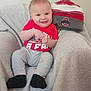 Austyn is registered to the contest to win money with this photo: baby, infant, smiling, red_shirt, gray_pants, black_socks, chair, blanket, hat, winter_hat, pom_pom, indoor, cute, happy, portrait, child, person, soft_texture, cozy, sports_fan