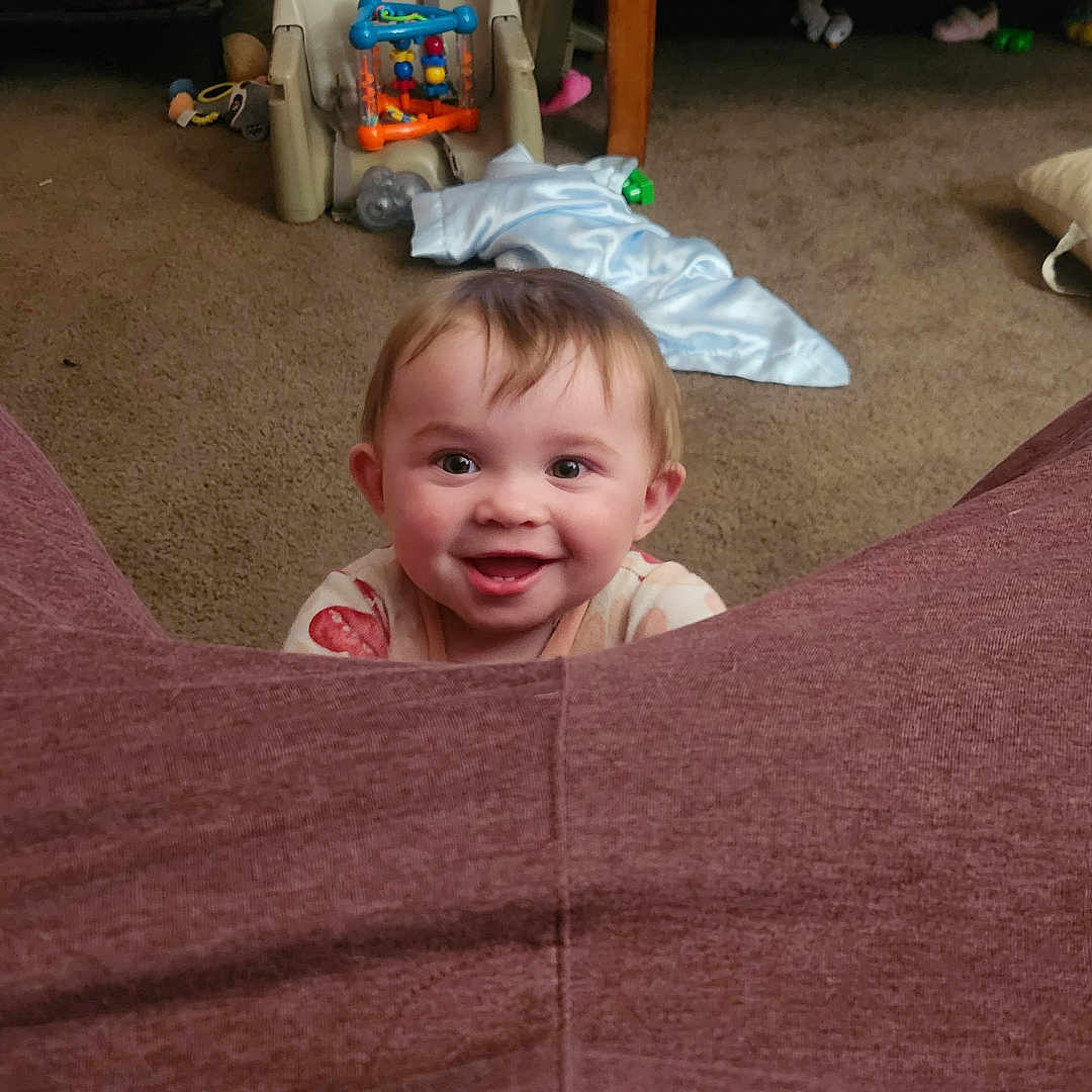 Emberlynn is registered to the contest to win money with this photo: baby, blanket, carpet, child, clothing, cup, face, floor, furniture, happy, highchair, home, indoor, living_room, person, play, seat, smiling, table, toy