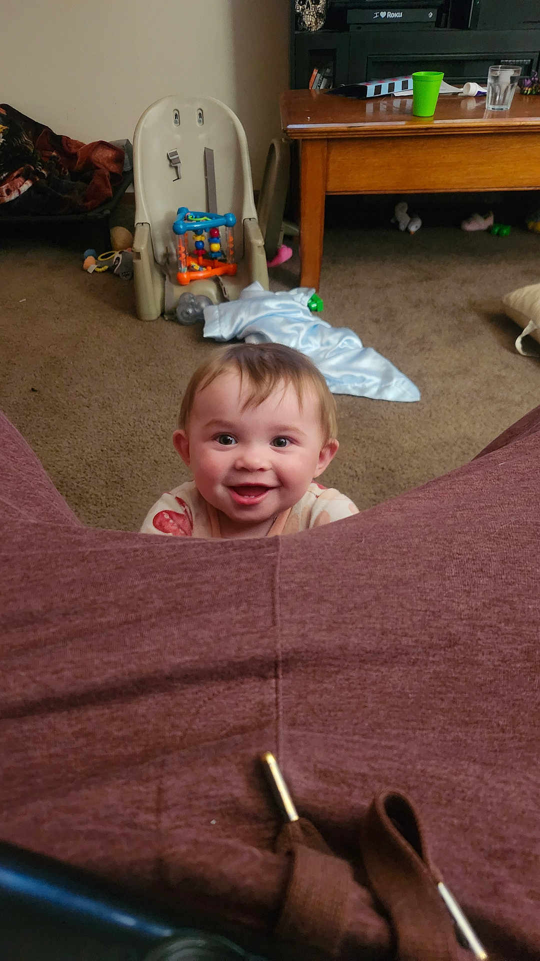 Emberlynn is registered to the contest to win money with this photo: baby, smiling, child, indoor, carpet, toy, highchair, table, cup, blanket, furniture, living_room, happy, person, face, floor, clothing, home, seat, play