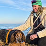 Baloo a rejoint le concours — aidez-le/la à gagner de superbes lots ! dog, person, beach, sea, sky, pebbles, green_beanie, jacket, sunglasses, leash, harness, petting, outdoor, casual_clothing, nature, daylight, calm, relaxation, companion, friendship