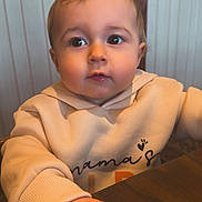 Raya is registered to the contest to win money with this photo: baby, child, toddler, face, hand, clothing, hoodie, hair, bow, table, indoor, restaurant, warm_lighting, cute, curious, portrait, person, sitting, wood, cozy