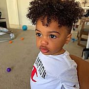 Justin is registered to the contest to win money with this photo: toddler, child, curly_hair, indoor, white_shirt, champion_logo, carpet, balls, toy, young_child, portrait, casual_clothing, expression, person, cute, curly_haircut, standing, room, furniture, gold_chain