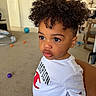 toddler, child, curly_hair, indoor, white_shirt, champion_logo, carpet, balls, toy, young_child, portrait, casual_clothing, expression, person, cute, curly_haircut, standing, room, furniture, gold_chain