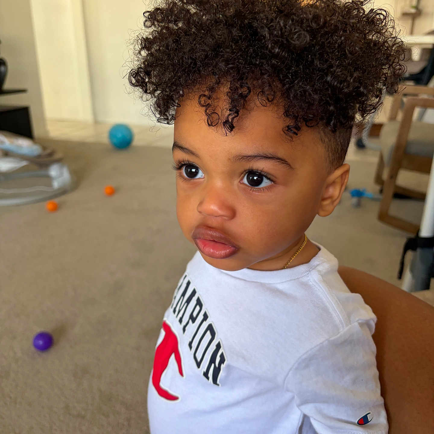 Justin is registered to the contest to win money with this photo: toddler, child, curly_hair, indoor, white_shirt, champion_logo, carpet, balls, toy, young_child, portrait, casual_clothing, expression, person, cute, curly_haircut, standing, room, furniture, gold_chain