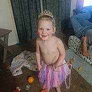 Athena joined the competition — help win amazing prizes! child, tutu, tiara, barefoot, toy, carpet, indoor, person, smile, happy, living_room, adult, television, curtains, pillow, clothing, floor, play, colorful, furniture