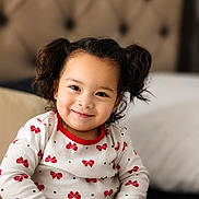 Ruth joined the competition — help win amazing prizes! child, toddler, girl, portrait, smiling, sitting, indoors, couch, blanket, curly_hair, hair_buns, brown_hair, patterned_clothing, red_bows, long_sleeves, earrings, face, happy, home, photography
