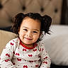 child, toddler, girl, portrait, smiling, sitting, indoors, couch, blanket, curly_hair, hair_buns, brown_hair, patterned_clothing, red_bows, long_sleeves, earrings, face, happy, home, photography