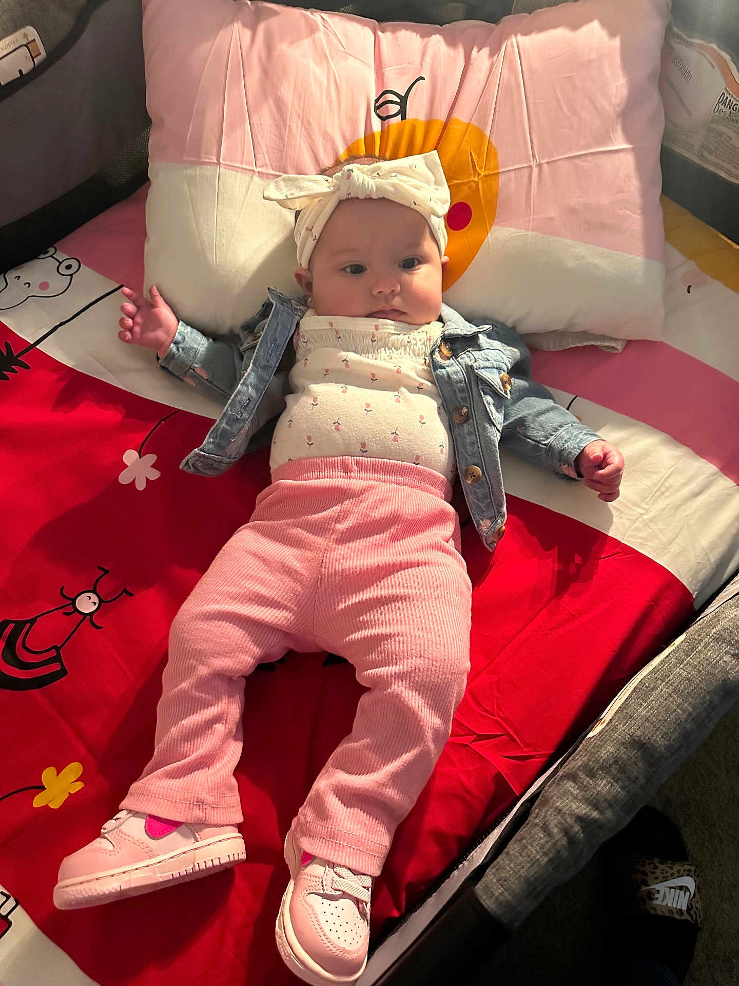 Jailyah joined the competition — help win amazing prizes! baby, infant, child, pink_pants, white_top, denim_jacket, headband, bow, sneakers, bed, pillow, cartoon_print, blanket, lying_down, indoor, cute, person, footwear, clothing, face