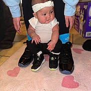 Jailyah is registered to the contest to win money with this photo: baby, child, headband, white_shirt, black_pants, black_shoes, small_shoes, adult_legs, hands, pink_rug, heart_pattern, floor_tiles, box, indoor, person, sitting, cute, footwear, home, cozy