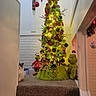 cat, christmas_tree, holiday, festive, grinch, plush_toys, christmas_lights, ornaments, indoor, staircase, carpet, home_decor, christmas_decorations, fluffy_cat, hallway, garland, santa_hat, green, red, cozy