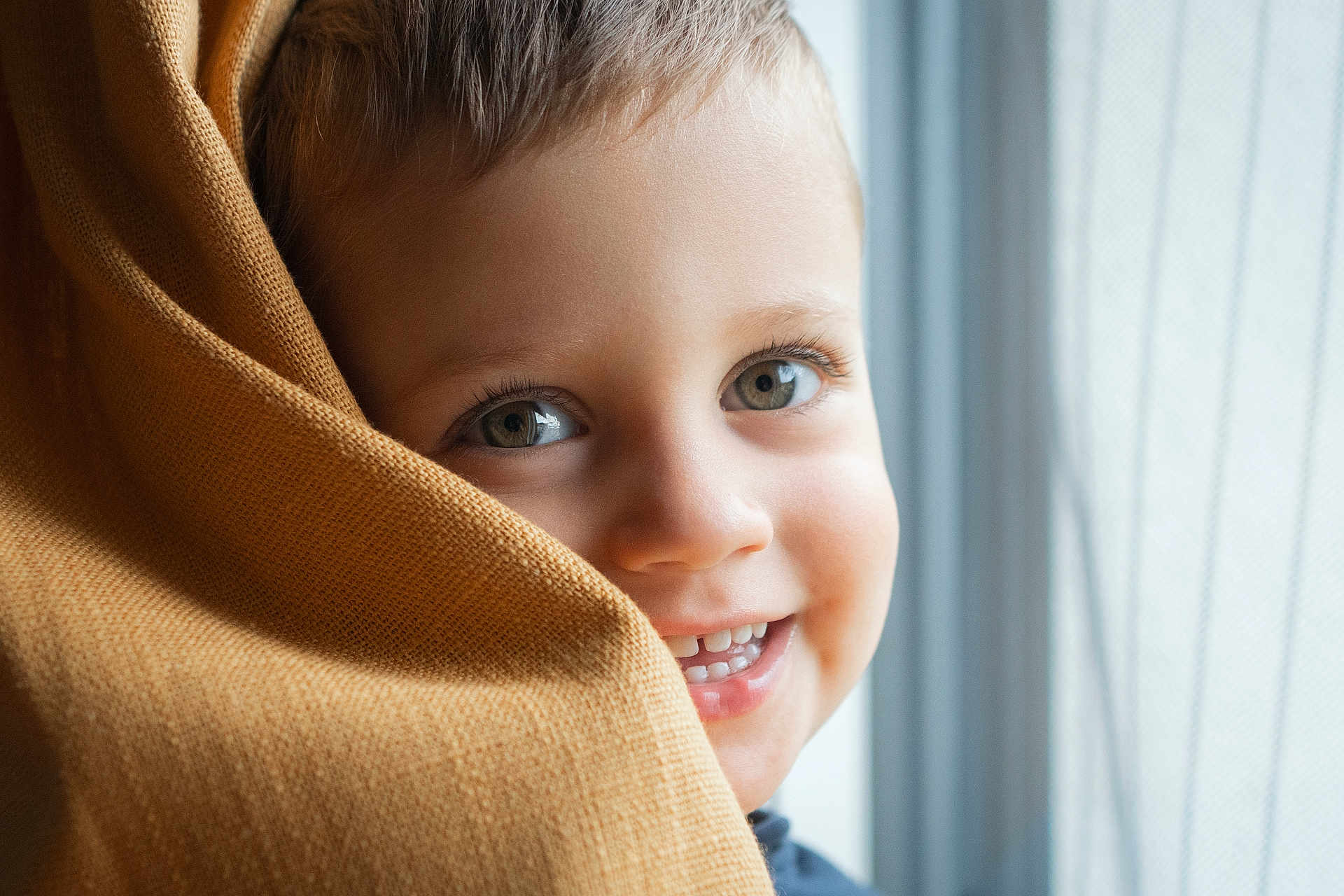 Elio participe au concours pour gagner de l'argent avec cette photo : toddler, child, smile, face, eyes, blanket, window, curtain, happy, close_up, portrait, indoors, cozy, soft_light, fabric, skin, cute, baby, person, young