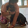 person, dog, indoor, carpet, hugging, pet, sunlight, glass_door, rocking_chair, floor, casual_clothing, short_hair, bracelet, paw, window, home, relaxation, affection, animal, comfort