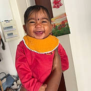 Alya a rejoint le concours — aidez-le/la à gagner de superbes lots ! baby, child, smiling, indoor, person, arm, clothing, red_outfit, orange_bib, face, happy, holding, painting, wall, door, earrings, home, wood_floor, furniture, socks