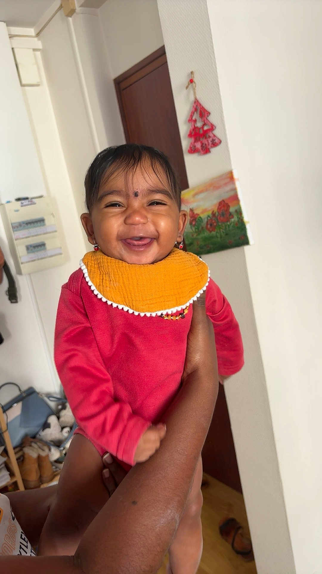 Alya a rejoint le concours — aidez-le/la à gagner de superbes lots ! baby, child, smiling, indoor, person, arm, clothing, red_outfit, orange_bib, face, happy, holding, painting, wall, door, earrings, home, wood_floor, furniture, socks