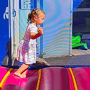 Noëline a rejoint le concours — aidez-le/la à gagner de superbes lots ! child, girl, dress, striped_clothing, barefoot, bounce_house, inflatable, play, outdoor, daylight, colorful, hair, braids, side_view, concrete, building, window, door, shadow, fun