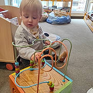 Alexandre participe au concours pour gagner de l'argent avec cette photo : toddler, child, toy, bead_maze, play, indoor, library, bookshelf, cushion, carpet, window, light, blond_hair, clothing, person, furniture, floor, hand, focus, young_child