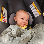 Cole is registered to the contest to win money with this photo: baby, car_seat, blanket, smile, infant, child, face, head, yellow_clothing, gray_blanket, warning_label, seat_belt, indoor, cute, portrait, person, cozy, young_child, happy, seated