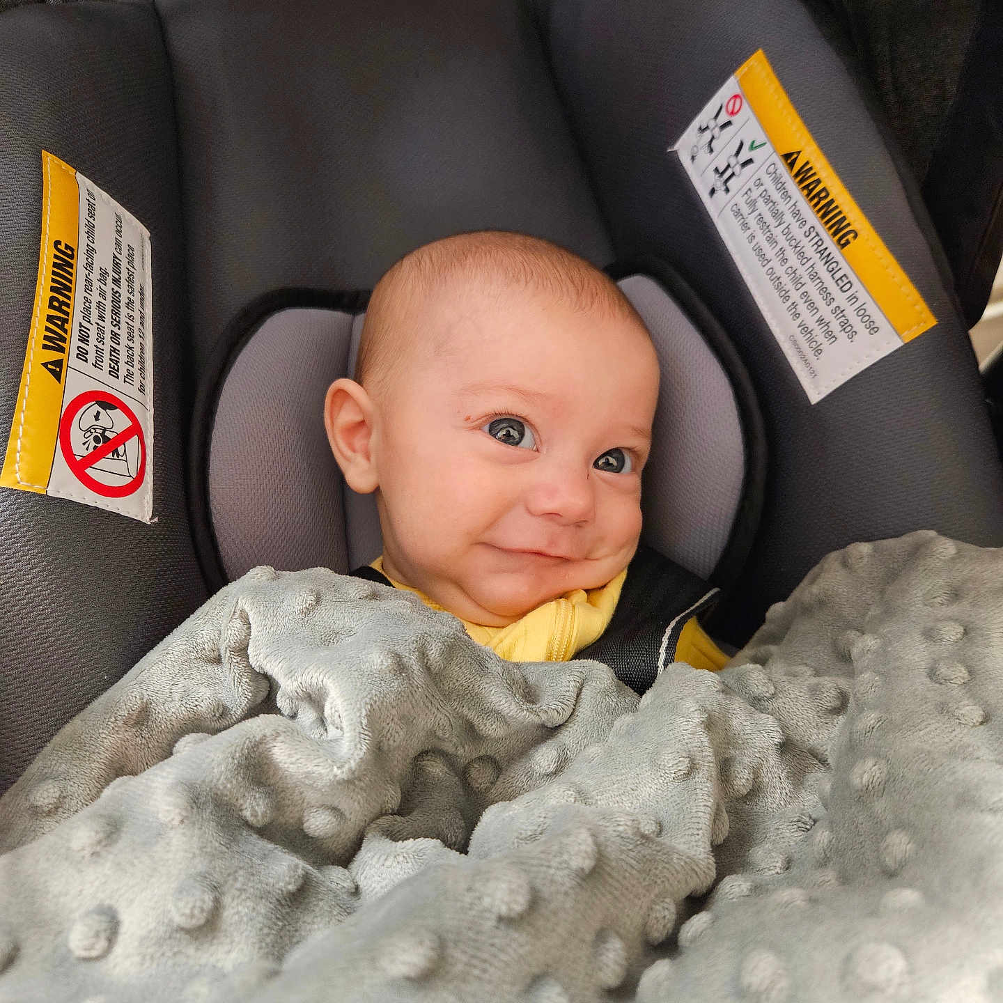 Cole is registered to the contest to win money with this photo: baby, blanket, car_seat, child, cozy, cute, face, gray_blanket, happy, head, indoor, infant, person, portrait, seat_belt, seated, smile, warning_label, yellow_clothing, young_child