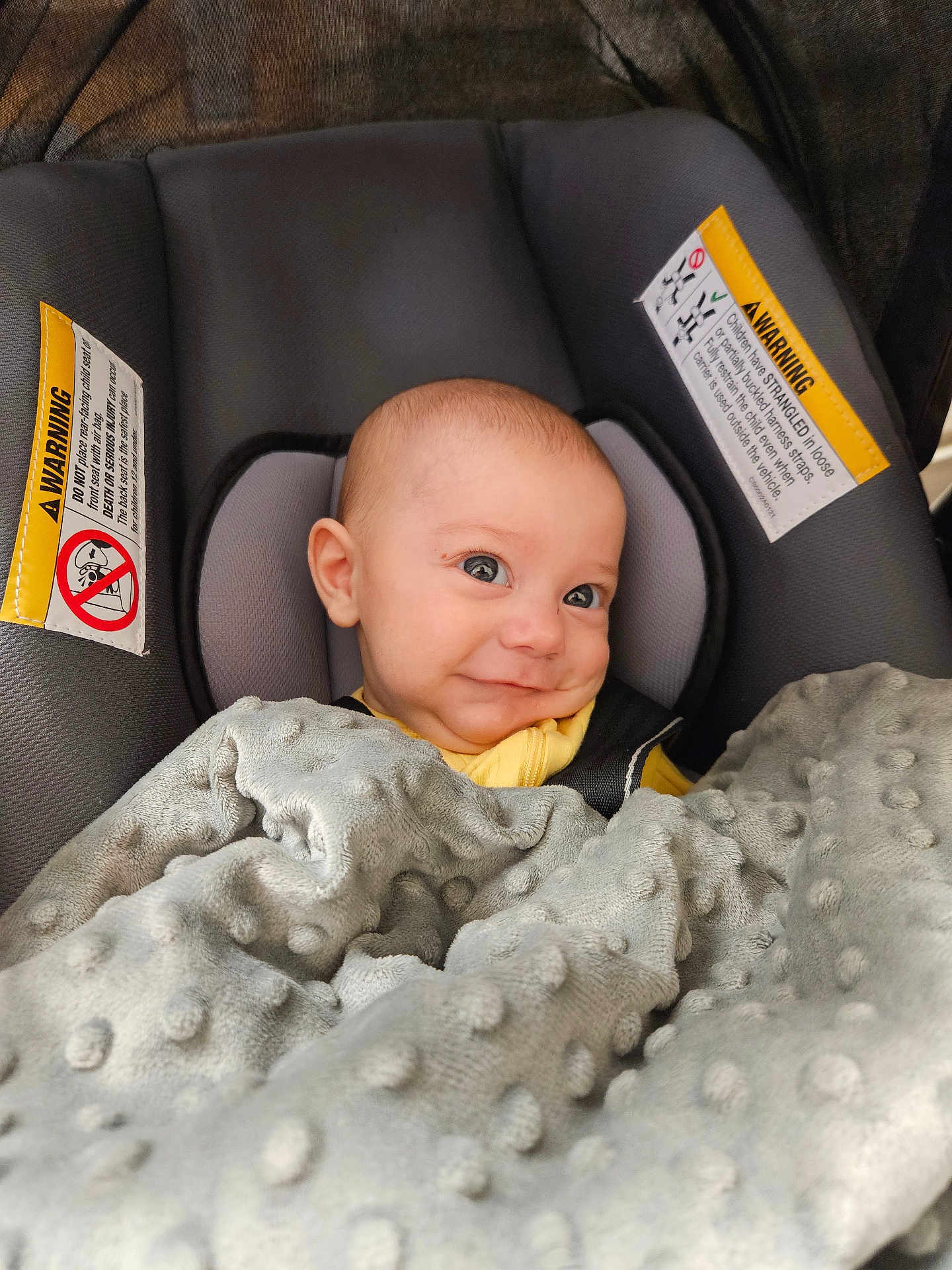 Cole is registered to the contest to win money with this photo: baby, car_seat, blanket, smile, infant, child, face, head, yellow_clothing, gray_blanket, warning_label, seat_belt, indoor, cute, portrait, person, cozy, young_child, happy, seated