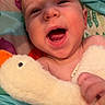 baby, laughing, plush_toy, duck, blanket, colorful, happy, infant, child, smiling, cute, soft_toy, cozy, bed, portrait, skin, hand, expression, comfort, indoors
