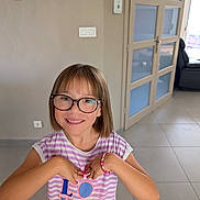 Louna a rejoint le concours — aidez-le/la à gagner de superbes lots ! child, girl, glasses, smiling, indoor, striped_shirt, bracelet, love_text, short_hair, happy, person, young, portrait, casual_clothing, modern_room, door, floor_tiles, natural_light, home, cute