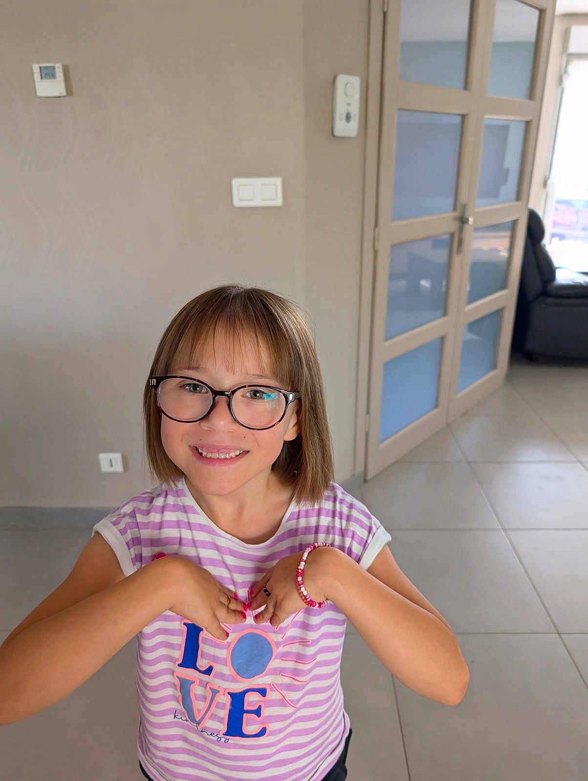 Louna a rejoint le concours — aidez-le/la à gagner de superbes lots ! child, girl, glasses, smiling, indoor, striped_shirt, bracelet, love_text, short_hair, happy, person, young, portrait, casual_clothing, modern_room, door, floor_tiles, natural_light, home, cute