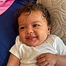 baby, child, infant, curly_hair, white_onesie, smiling, lying_down, bed, blue_sheet, beige_pillow, quilted_pillow, hand, earring, cute, happy, indoors, portrait, skin, face, person