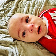 Julius joined the competition — help win amazing prizes! baby, infant, face, eyes, mouth, blanket, bed, clothing, red, white_trim, skin, head, expression, indoors, portrait, closeup, person, cute, lying_down, soft